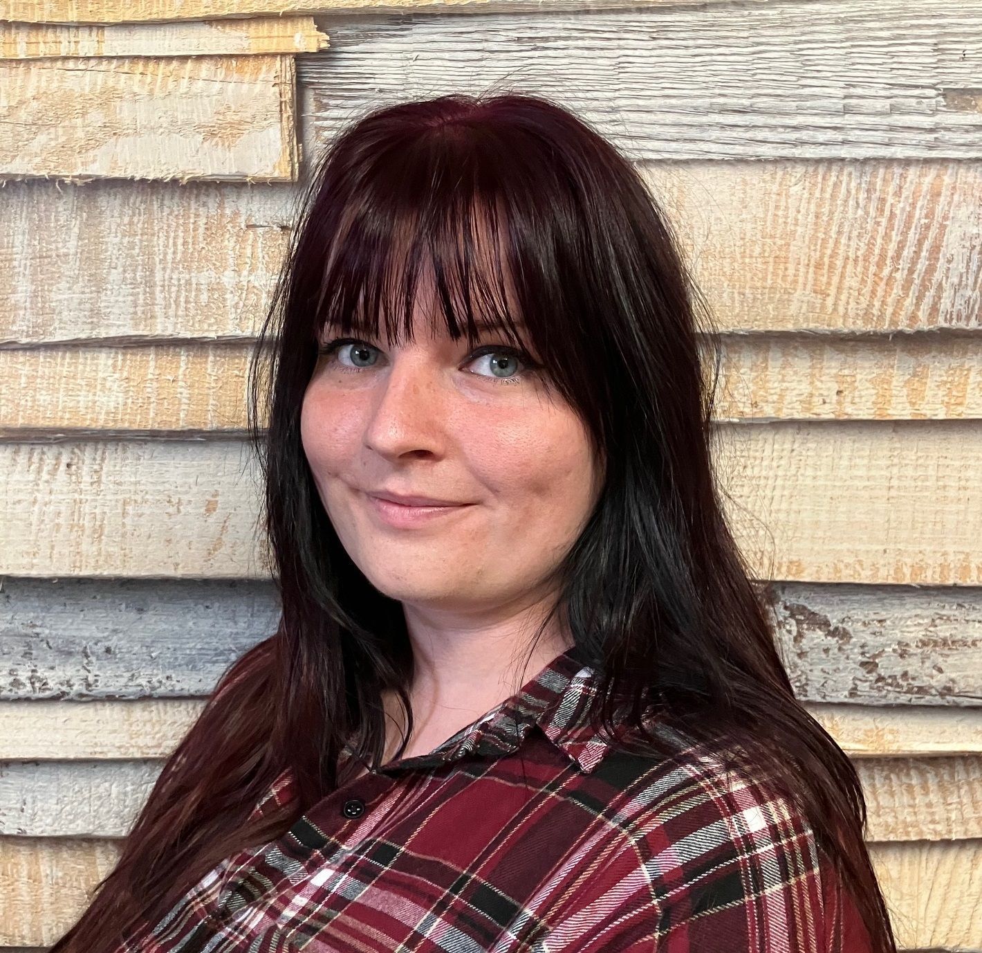 Frau mit dunklen Haaren in einem rotkarierten Hemd posiert vor einer rustikalen Holzwand