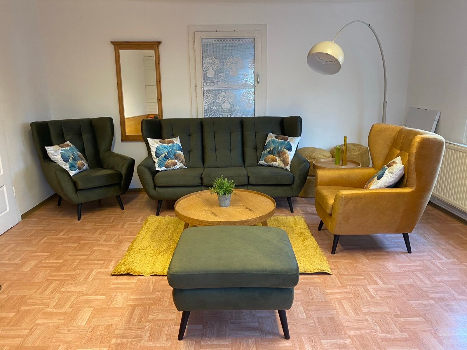 Gemütliches Wohnzimmer mit grünem Sofa und Sesseln, gelbem Teppich, Hocker, Stehlampe und Wandspiegel.