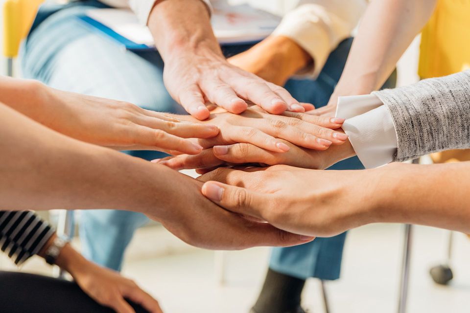 In einem Innenraum, in dem Menschen um einen Tisch sitzen, sind die Hände zu einer Geste der Teamarbeit zusammengelegt.