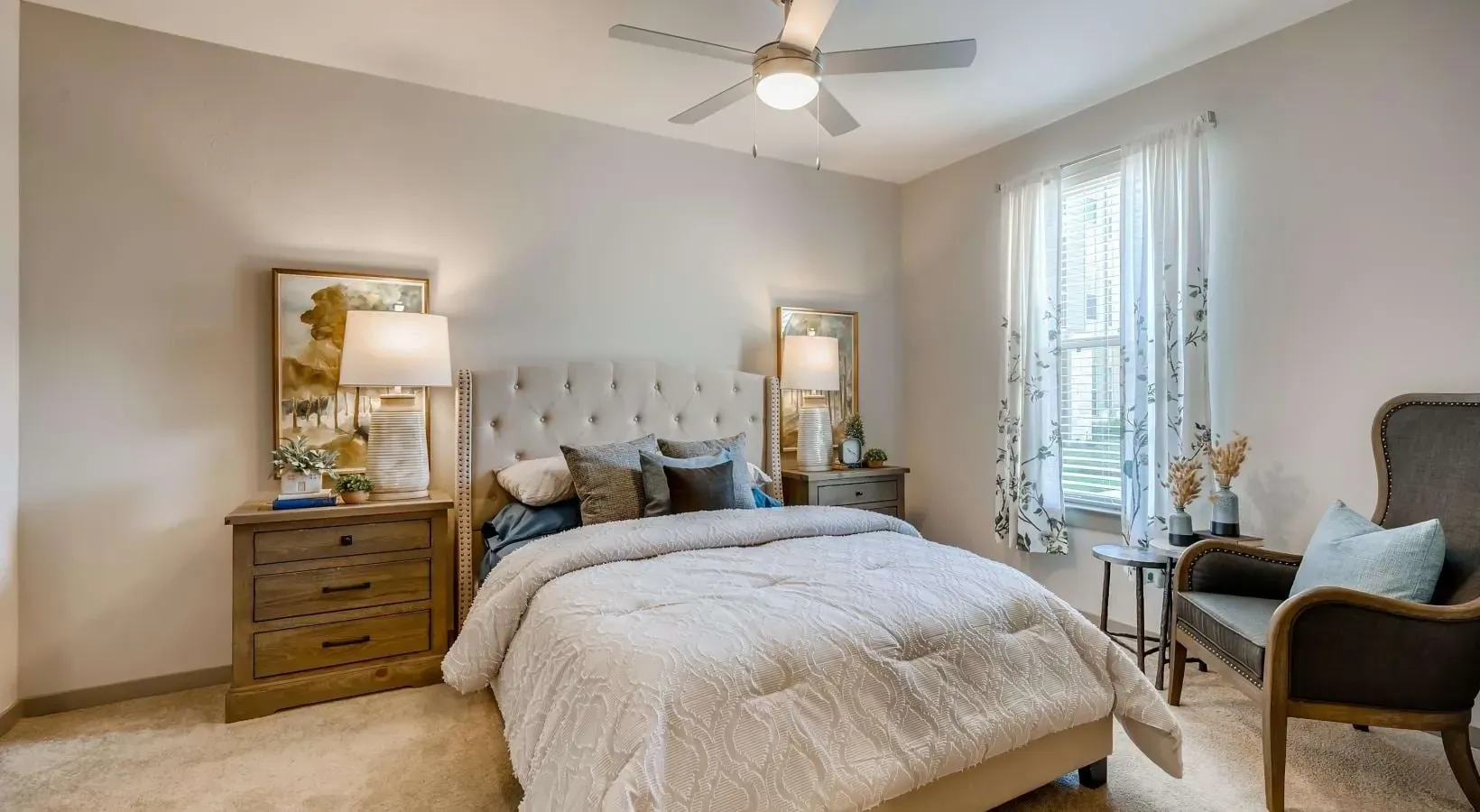 Bedroom scene with tufted headboard, two wooden nightstands, lamps, and a window with sheer floral curtains.