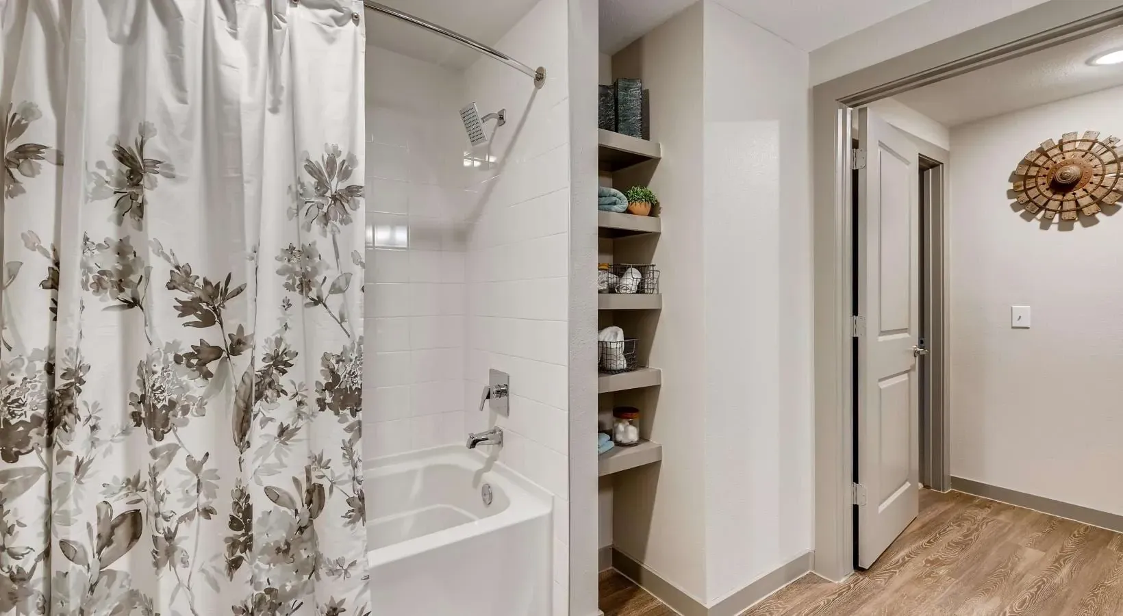 Bathroom in apartment with tub/shower, white tiles, floral shower curtain, and open shelving.