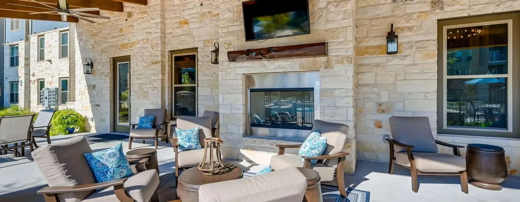 Outdoor patio with stone wall, fireplace, mounted TV, and cushioned lounge chairs.