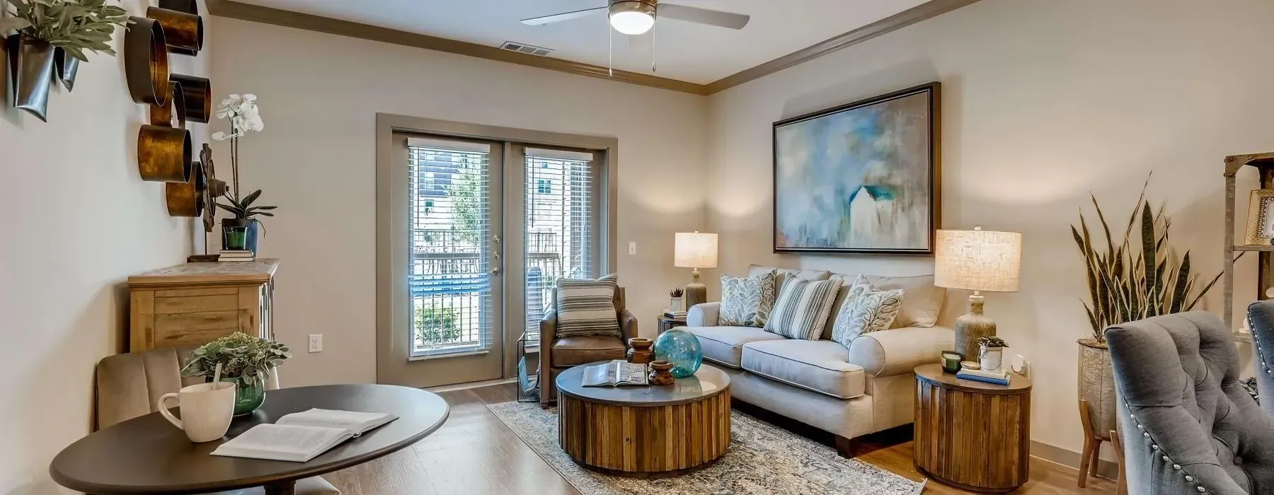 Bright apartment living room with beige sofa, armchair, and two round wooden tables beside a glass door.