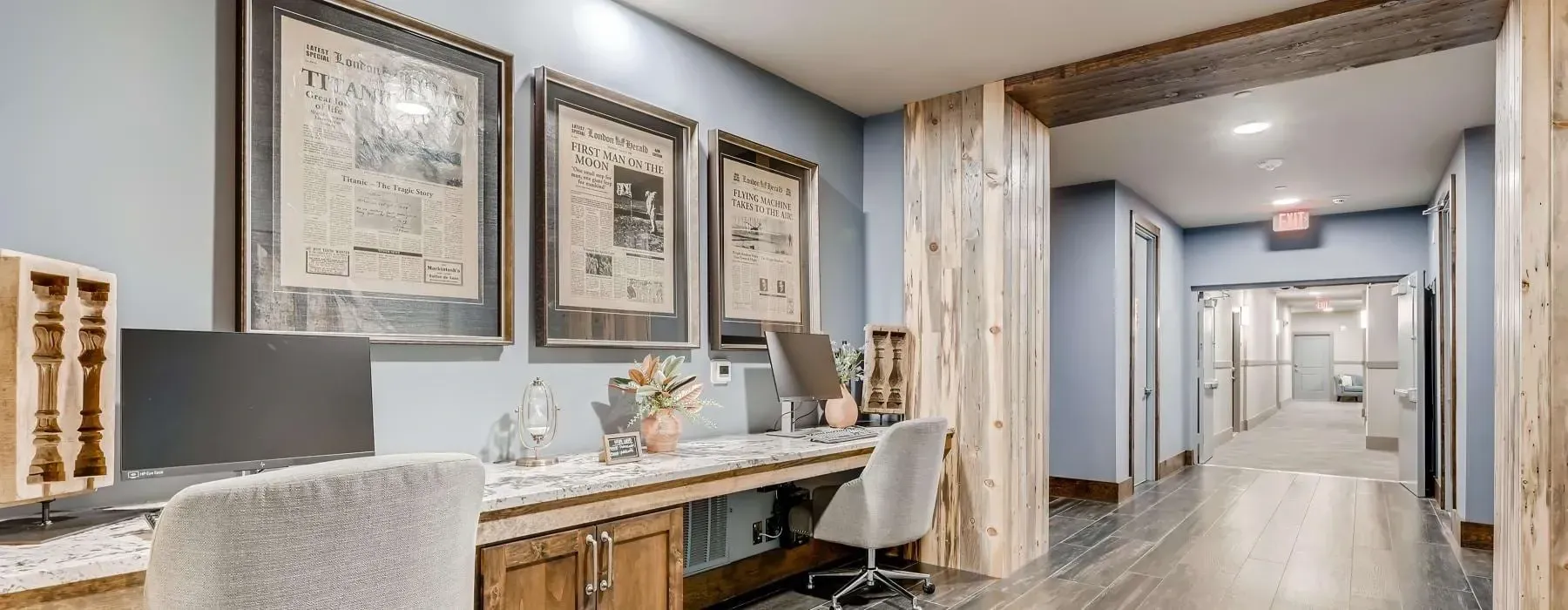 Interior apartment lobby with a long desk, two computer monitors, framed wall art, and a hallway.