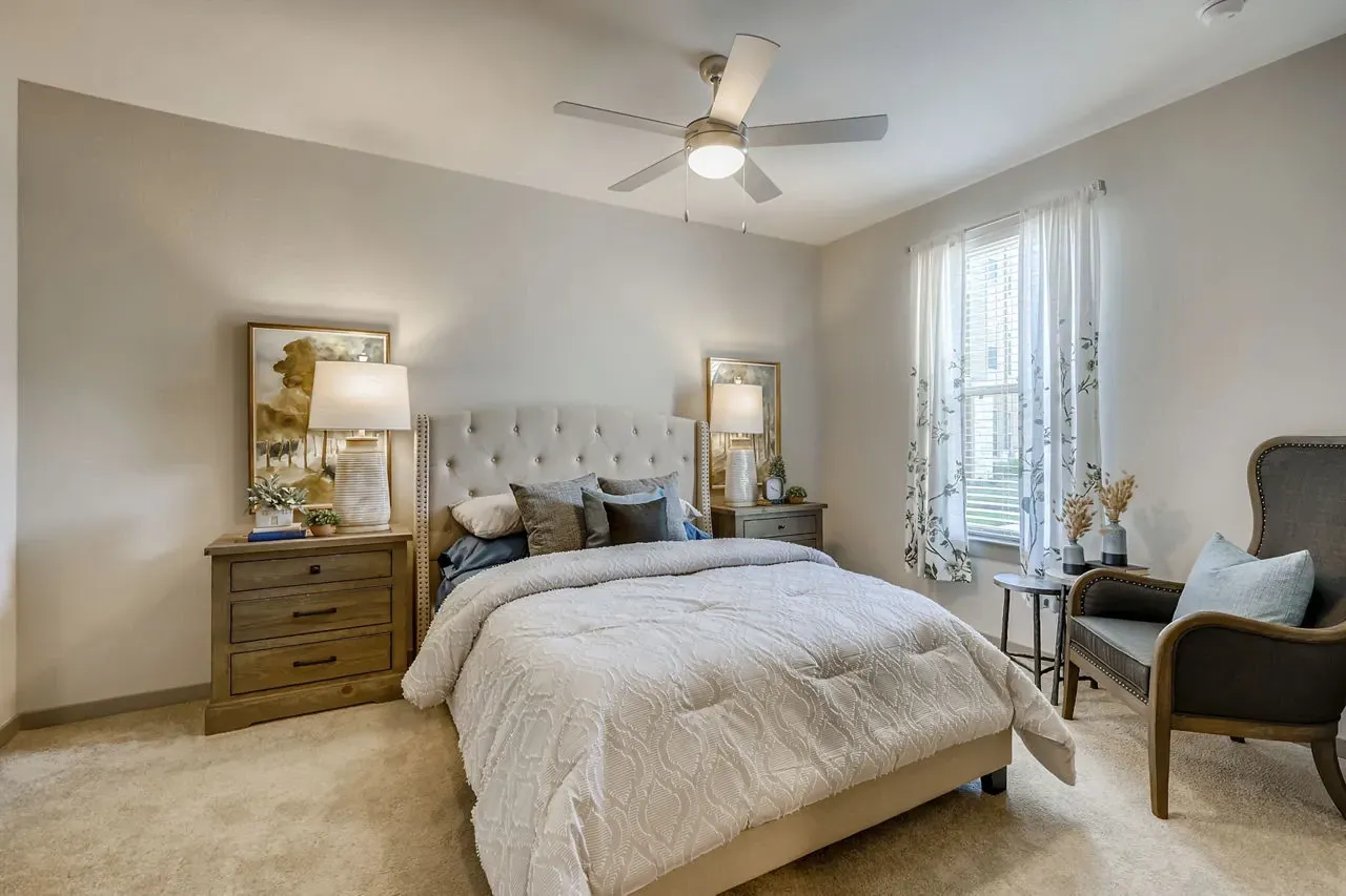 Bedroom in an apartment: beige tufted bed, two nightstands, lamps, and a chair by a window.