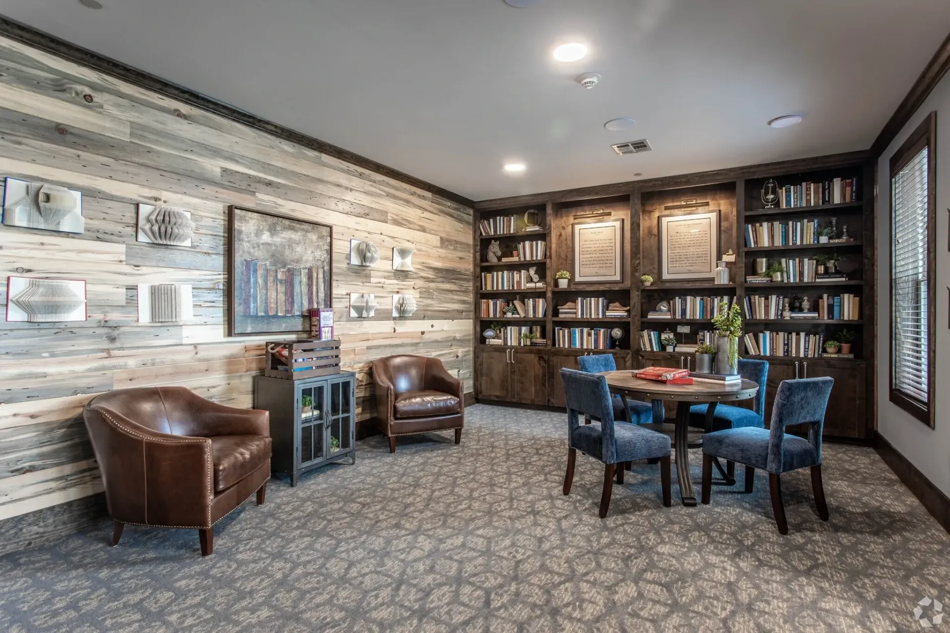 Interior community library/lounge with wood plank accent wall, built-in bookshelves, and seating around a round table.