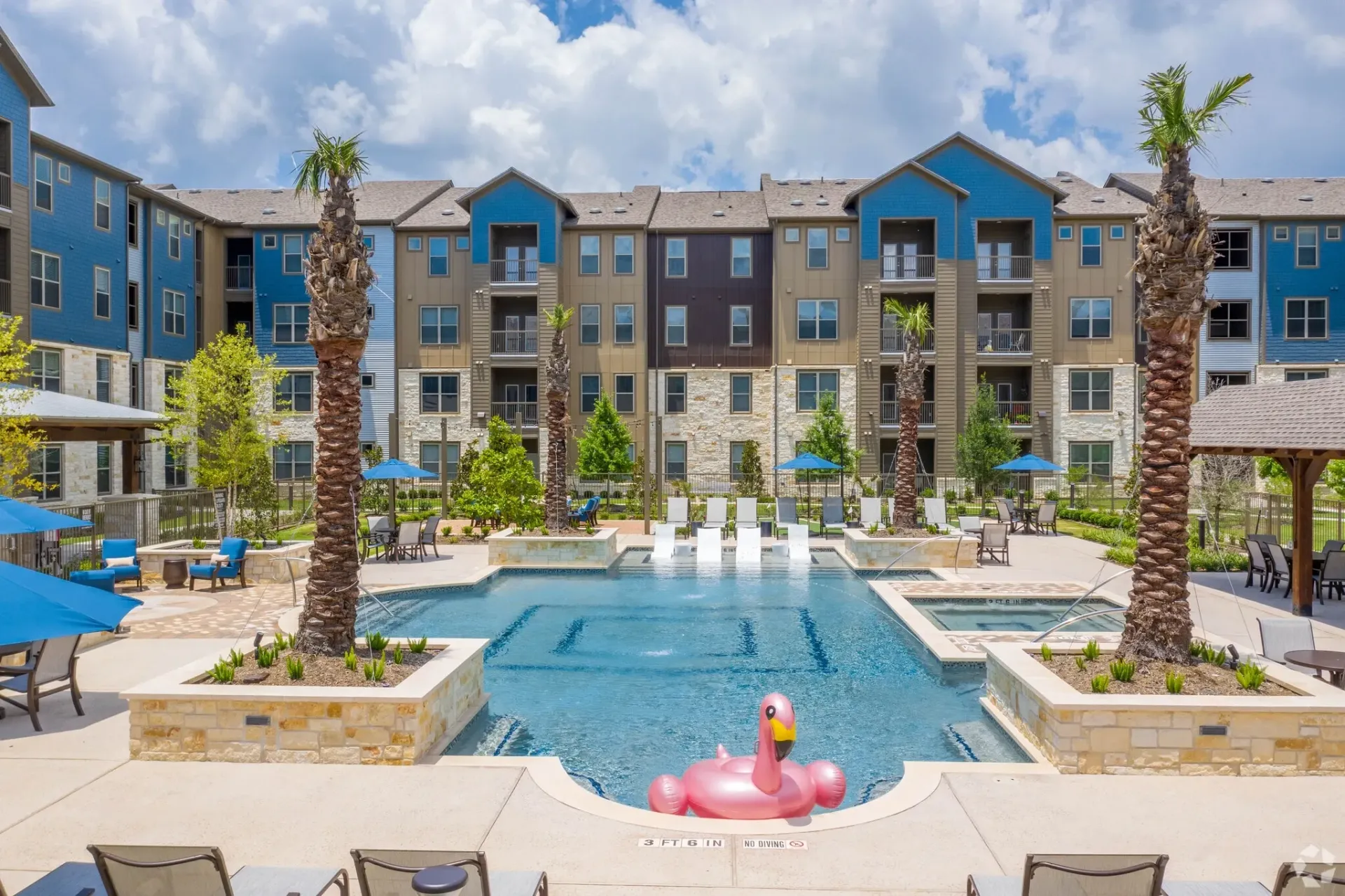 Outdoor pool area at a modern apartment community with palm trees, lounge chairs, and umbrellas.