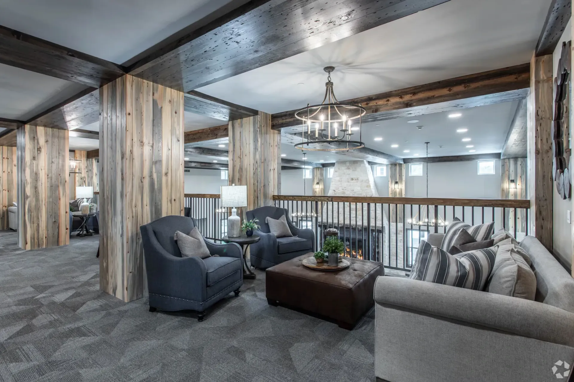 Rustic indoor resident lounge with timber columns, gray seating, a stone fireplace, and a circular chandelier.