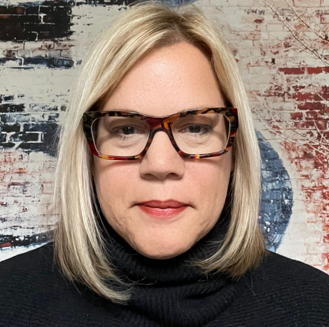 Woman with blonde hair and glasses wearing a black turtleneck in front of a brick and painted wall.