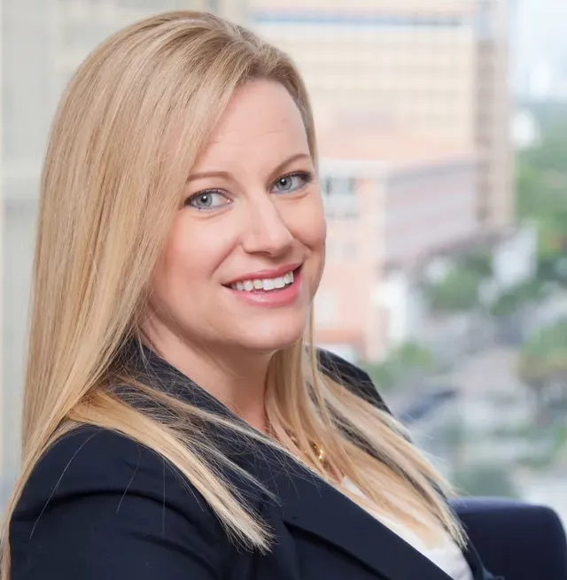 Blonde woman in a navy blazer smiles at the camera, city backdrop.