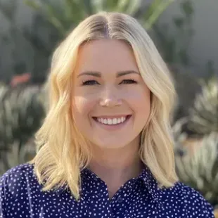Blond woman smiling, wearing a blue polka-dot shirt, with a background of greenery.