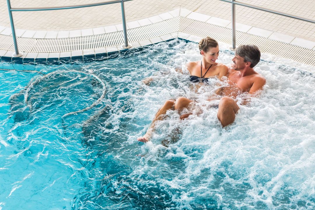 Paar im Whirlpool, Wasser plätschert. Sie lächeln und schauen sich in einem Hallenbad an.