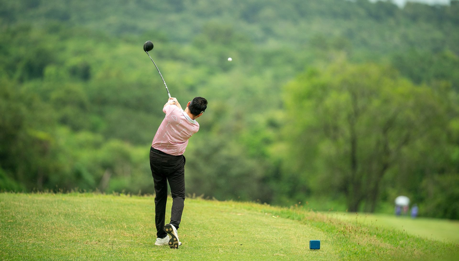 Ein Golfer schwingt den Schläger auf dem Grün, der Ball fliegt in der Luft. Üppige grüne Landschaft, Bäume im Hintergrund.