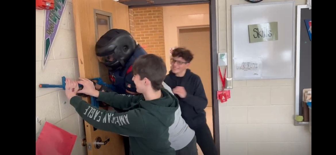 A person in tactical gear holding a blue rifle enters a hallway where people are lined up against the wall, showing expressions of fear.