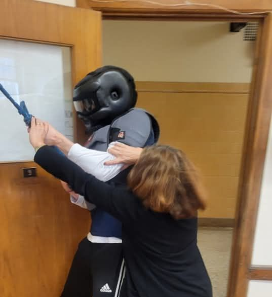 Two men practicing combat in a gym. One in civilian clothes with a handgun, facing another in a blue uniform and headgear.