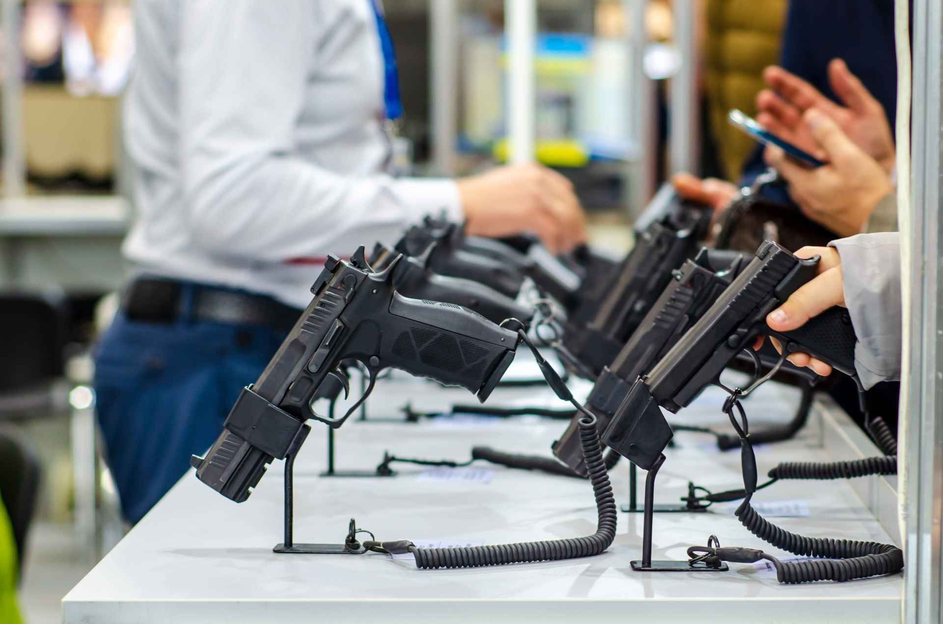 Guns displayed on a table at a gun show; people browse and handle the firearms.