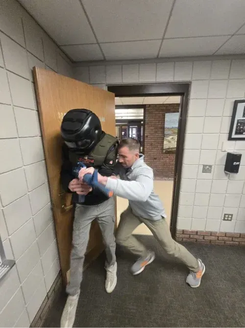 A person in tactical gear is pushed against a doorway by another person in a building hallway.