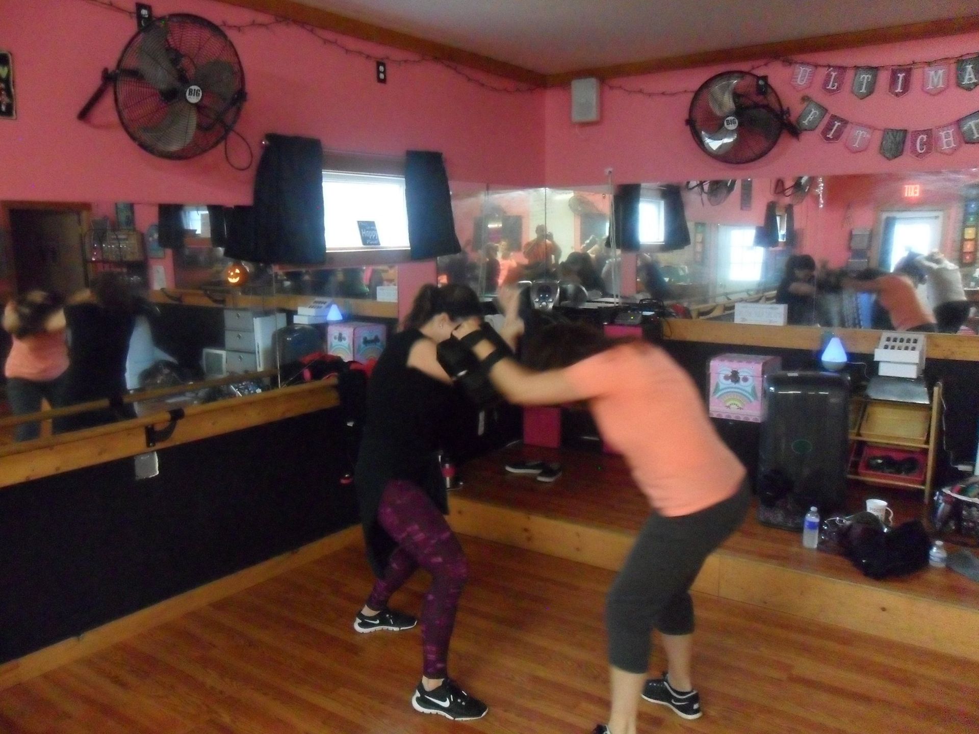 Two women in a pink gym spar, one throwing a punch, other blocking. Room has fans, mirrors, and other fitness equipment.