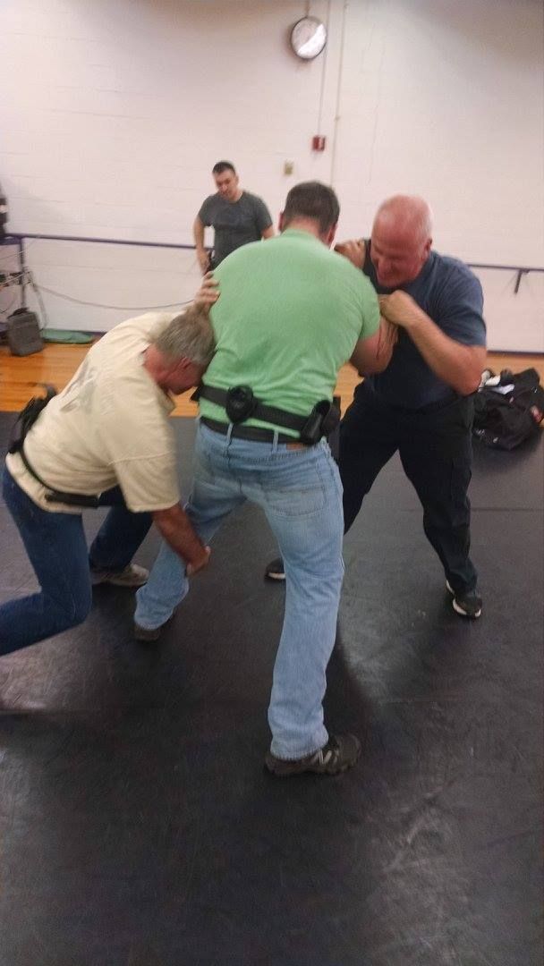 Three men in a gym practice a self-defense technique. One man is held between the other two, who are practicing a grappling move.