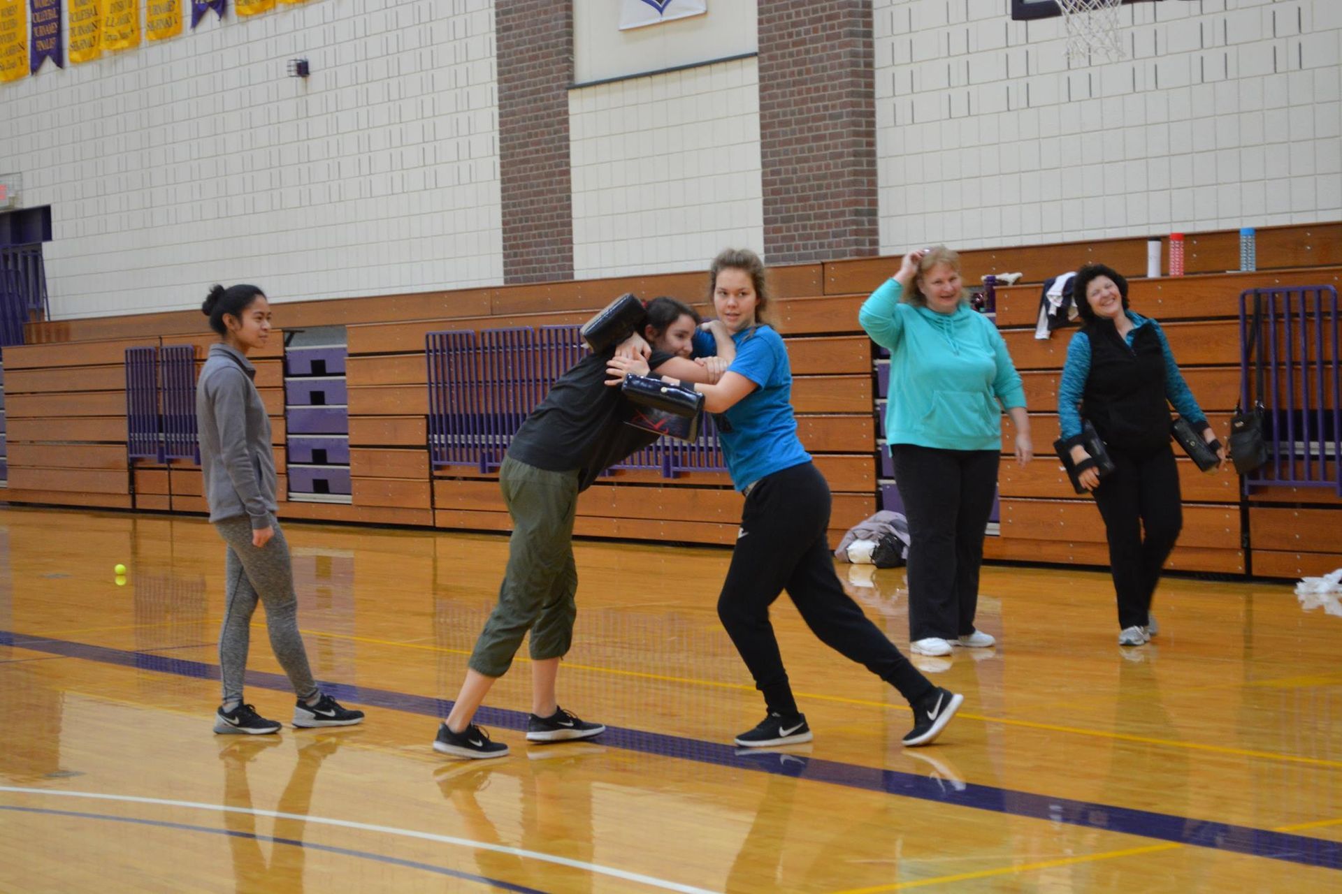 People practicing self-defense in a gym. Two women are grappling, others observe. The gym has a wooden floor and purple accents.