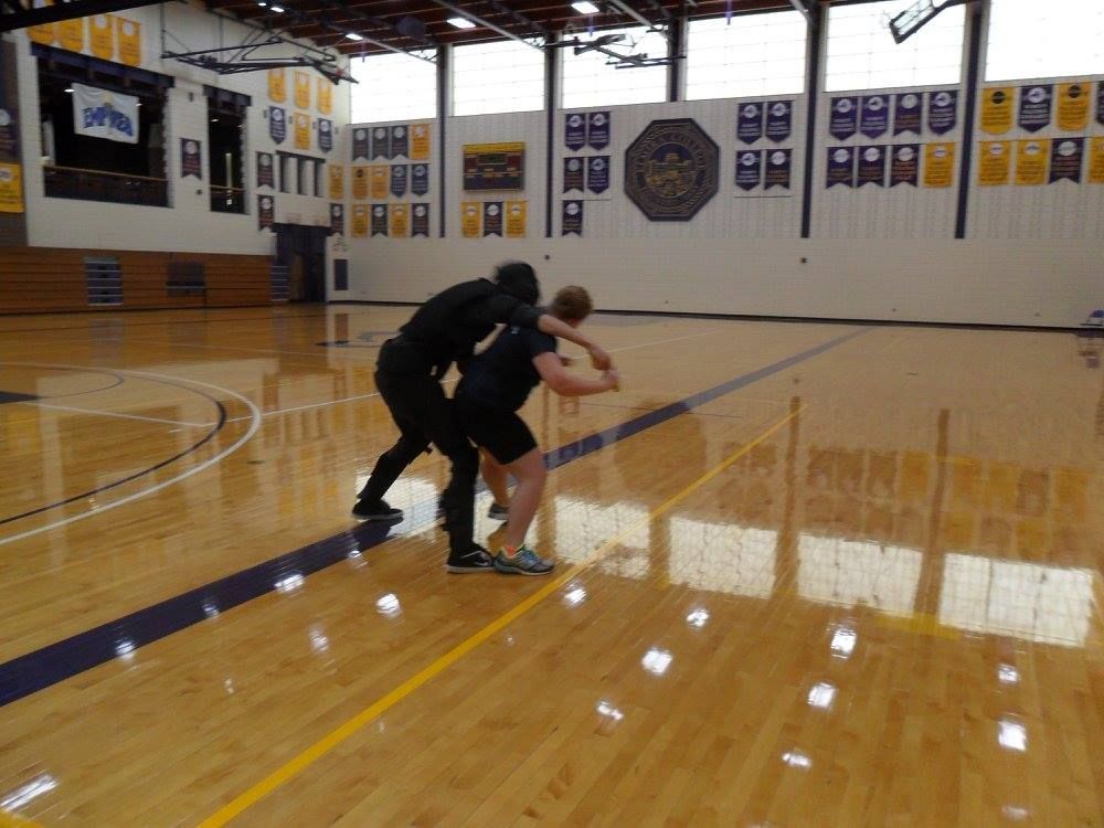 Two people, wearing black clothing, are wrestling in a gymnasium. They are on a shiny wooden floor with blue and yellow lines.