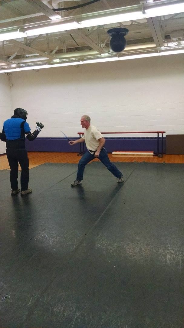 Man in white shirt practices fencing with a person wearing protective gear on a black mat indoors.