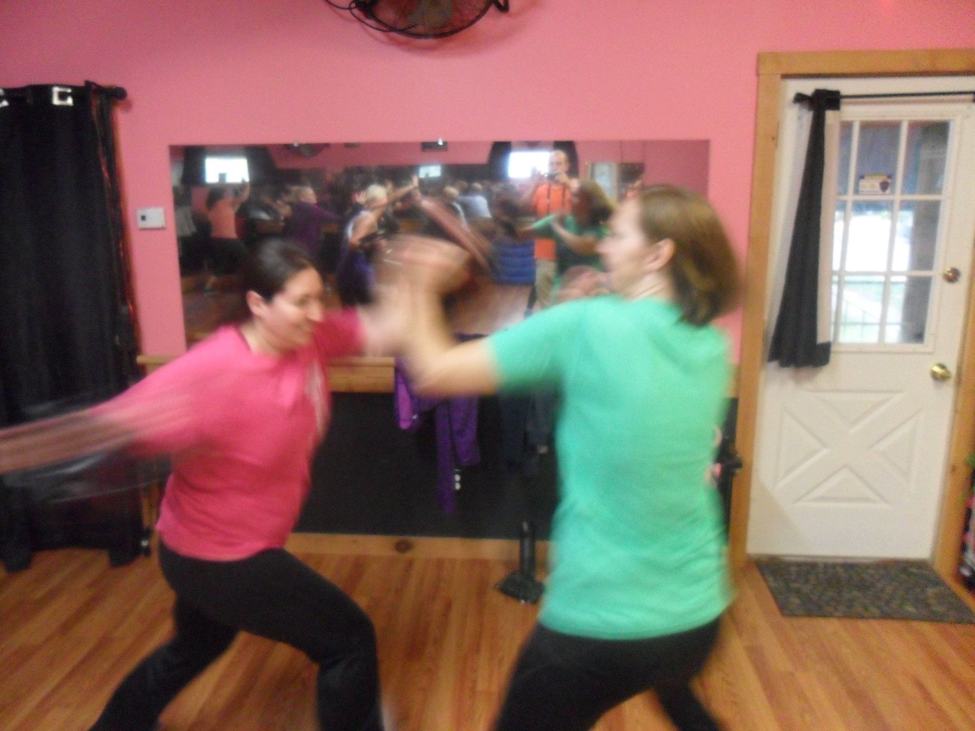 Two women in a dance studio, one in pink striking at the other in green, both with blurred motion.