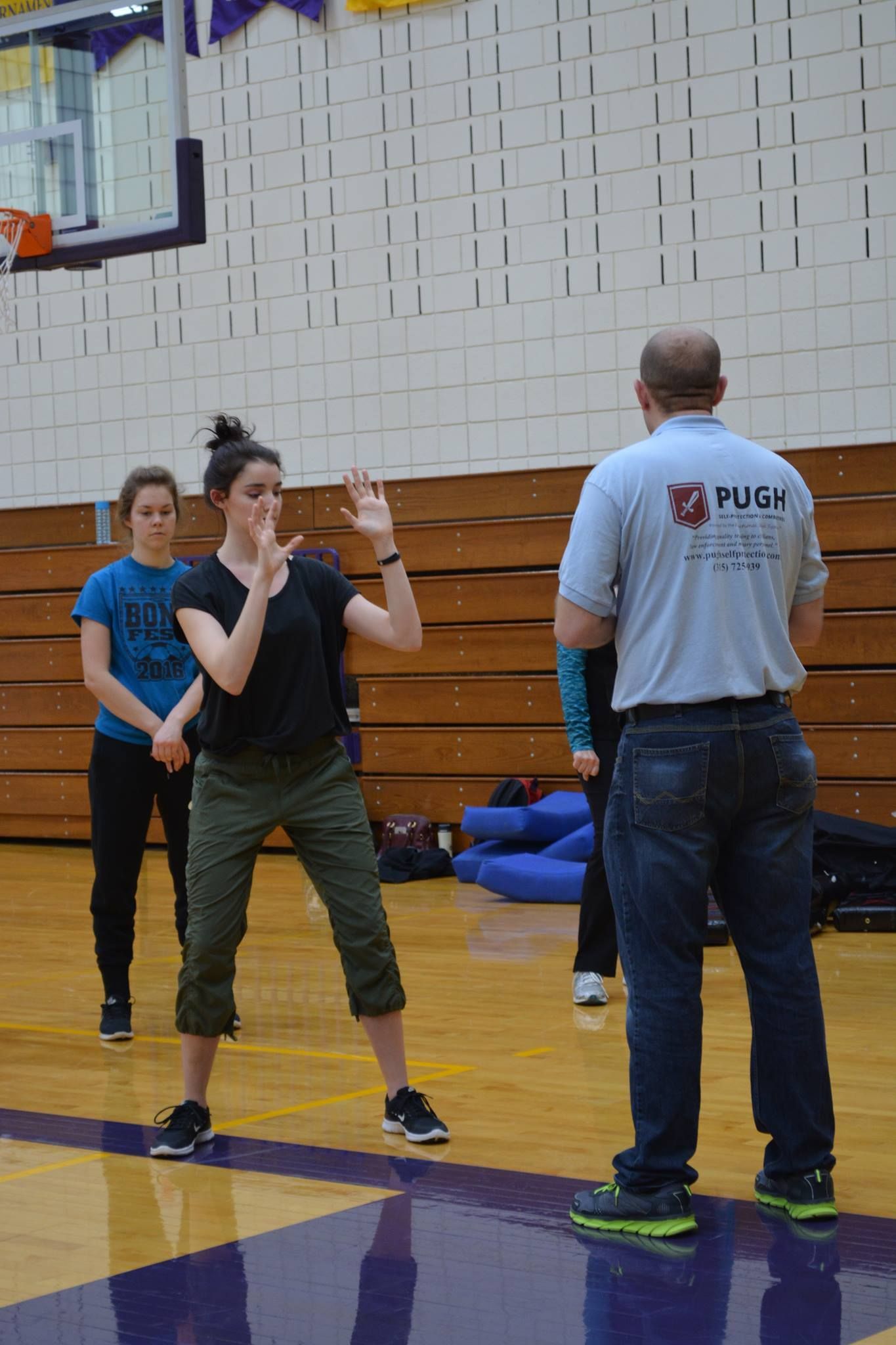 A woman in a black shirt and green pants practices self-defense with a bald instructor in a gym. Other people watch.