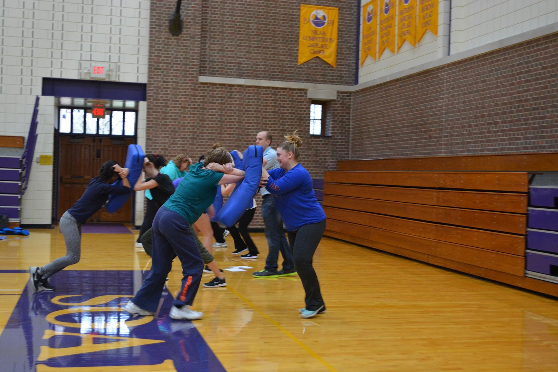 People in a gym practice self-defense techniques, kicking and punching blue pads. The setting has purple and gold accents.