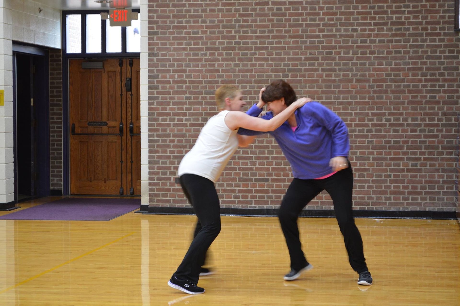 Two women wrestling in a gym with a wooden floor and brick wall. One wears white, and the other wears purple, both with athletic wear.