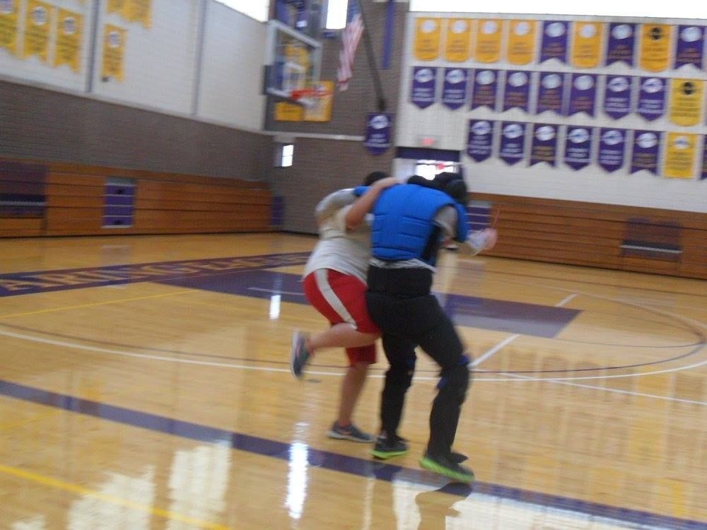 Two people wrestling in a gymnasium; one wears a blue vest, black pants and the other, a white shirt and red shorts.
