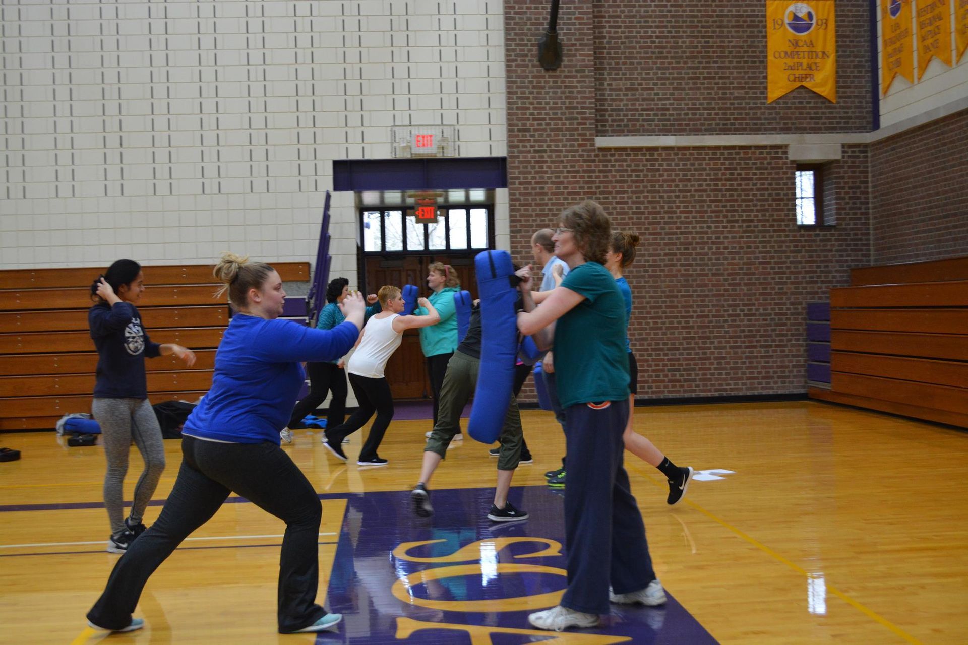 People in a gym practice self-defense with padded batons. Several women, some wearing blue, are shown striking and blocking.