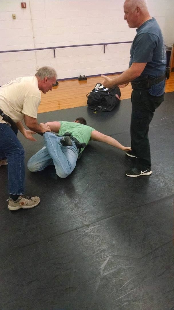 Three men demonstrating a takedown on a padded floor; one man on the ground is being held by the other two.