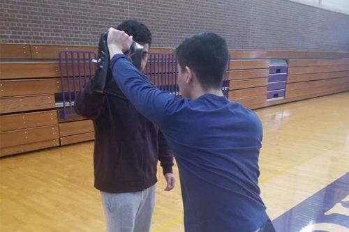 Two people practice fencing in a gymnasium. One person holds a fencing mask up near the other’s head.