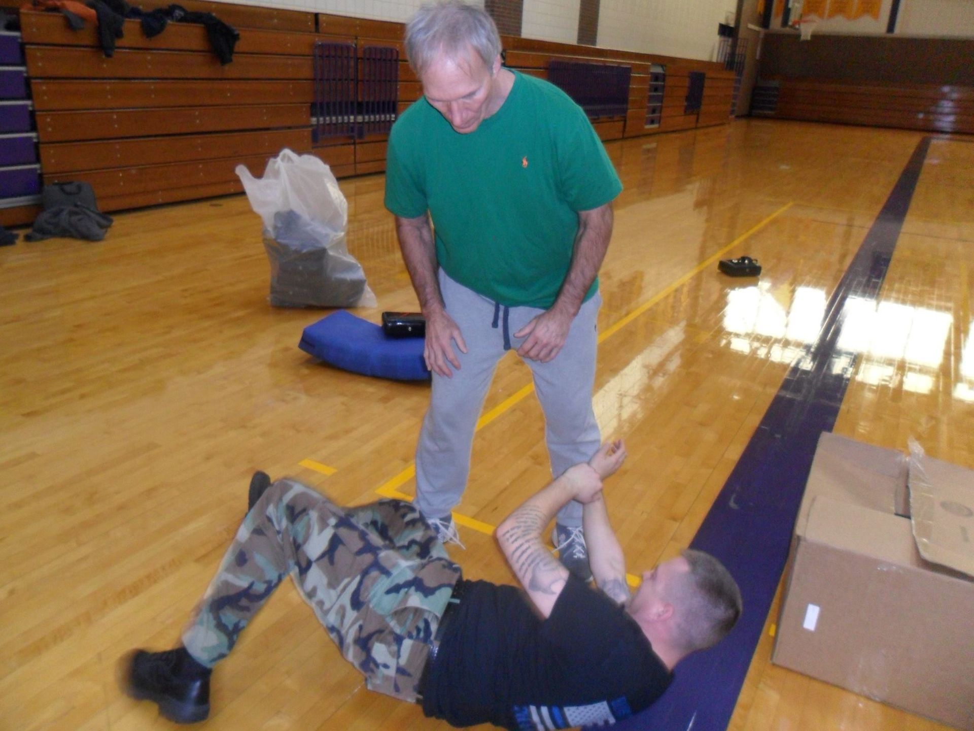 Man in green shirt assisting another man in camouflage pants on a gym floor. The man on the floor is holding his arm as if injured.