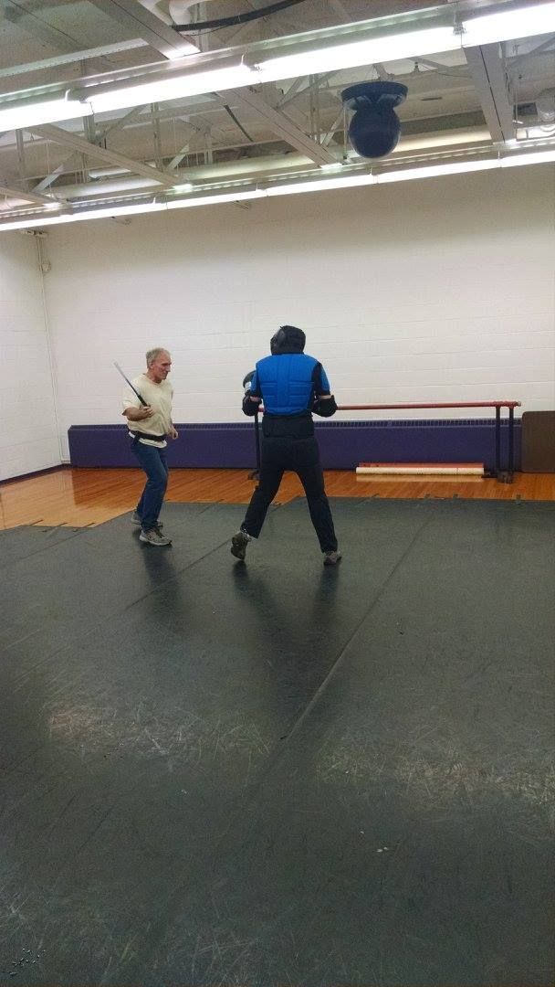 Two men sparring with swords in a gym. One in protective gear, facing the other who holds a sword and wears casual clothes.