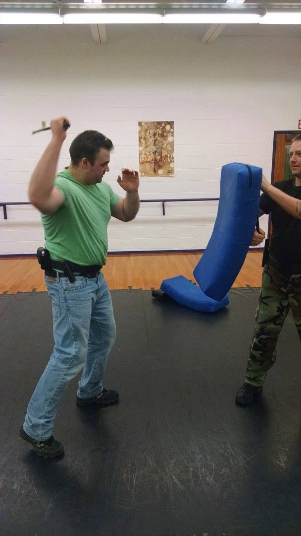 Man in green shirt practices knife defense against a target held by another person. They're in a gym.