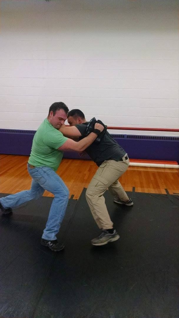 Two men in a wrestling stance on a mat. One in green shirt lunges forward, attempting to grab the other in black. Gymnasium setting.
