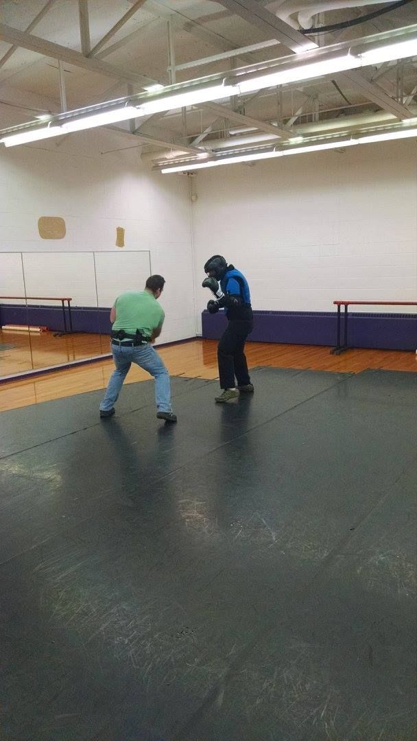 Two men practicing combat in an indoor gym. One in a blue uniform and protective gear faces the other in a green shirt and jeans, both on a padded mat.