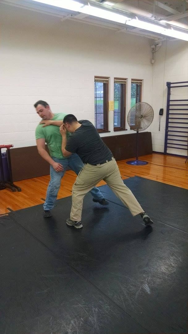 Two men practice self-defense on a black mat. One chokes the other, who is wearing a green shirt, while the other man blocks.