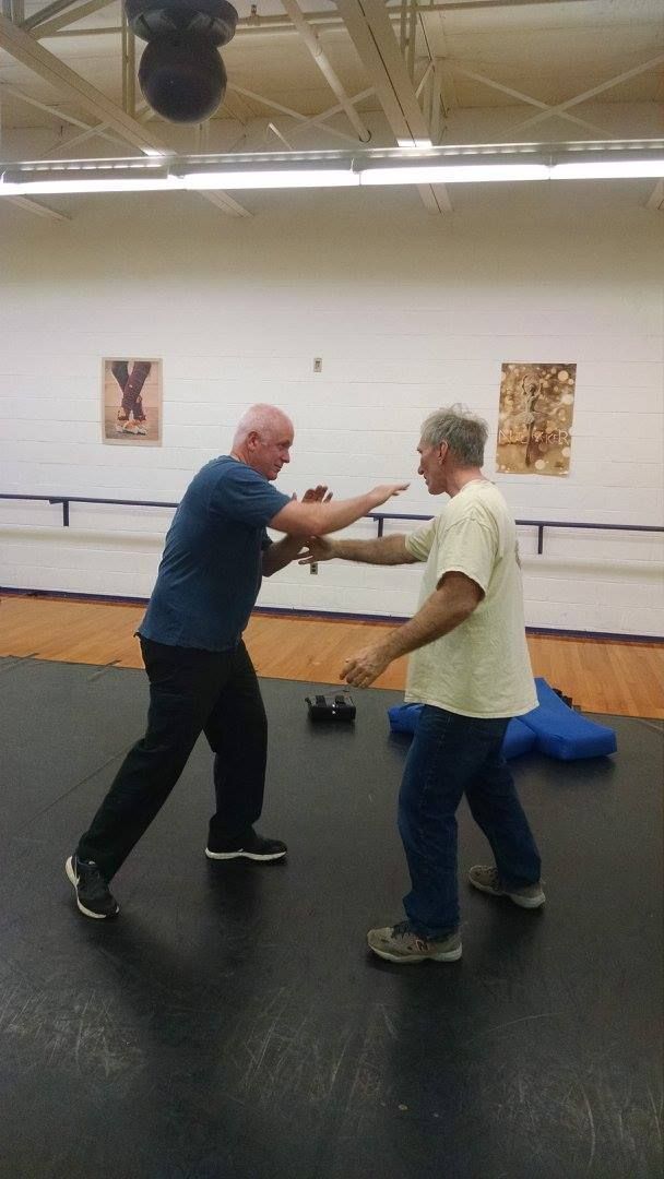 Two men practicing martial arts in a gym. One blocks a punch from the other, who wears a blue shirt and black pants.