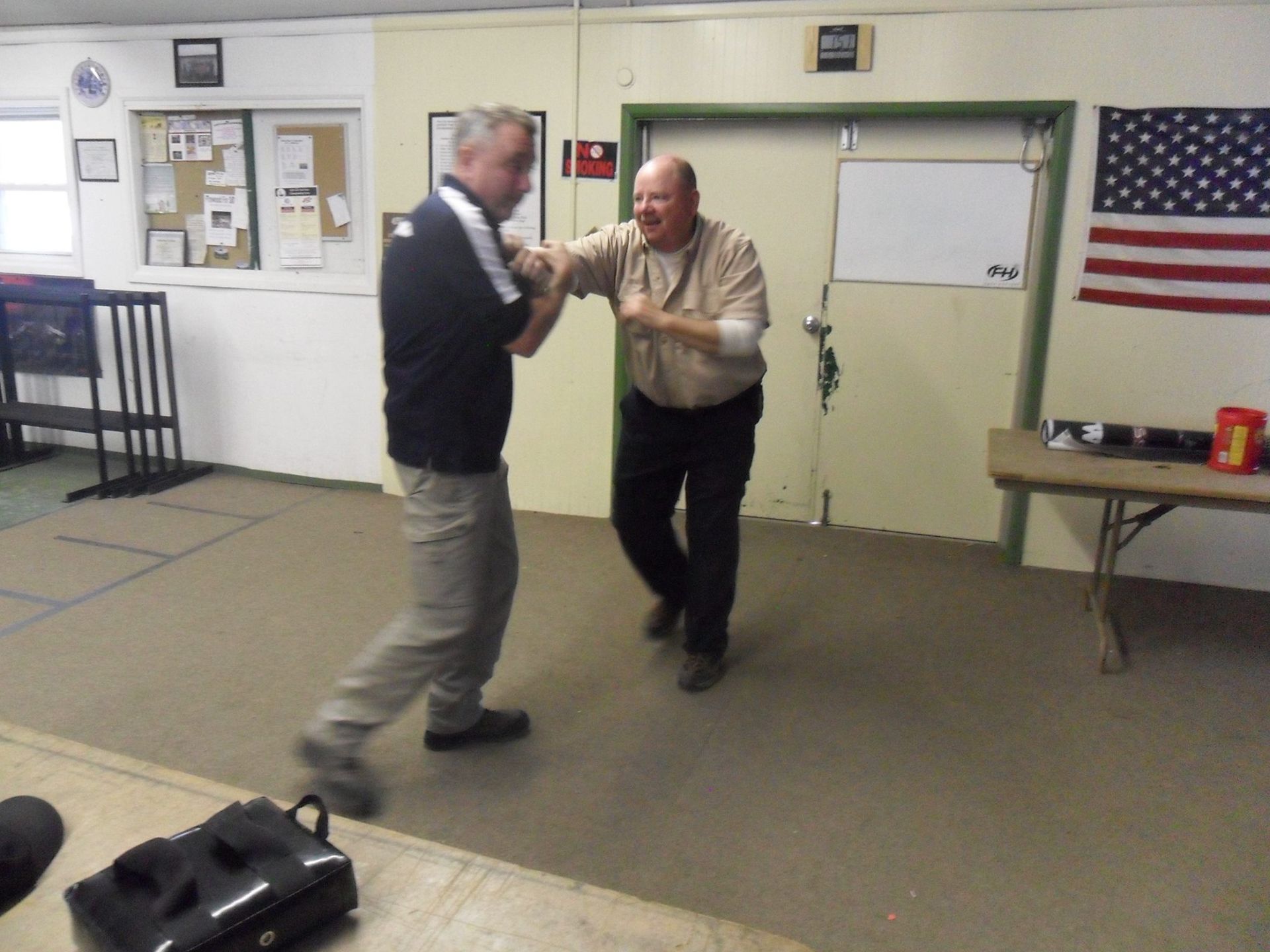 Two men in a room, one throwing a punch, possibly a self-defense demonstration. The setting is a plain room with an American flag and a table.