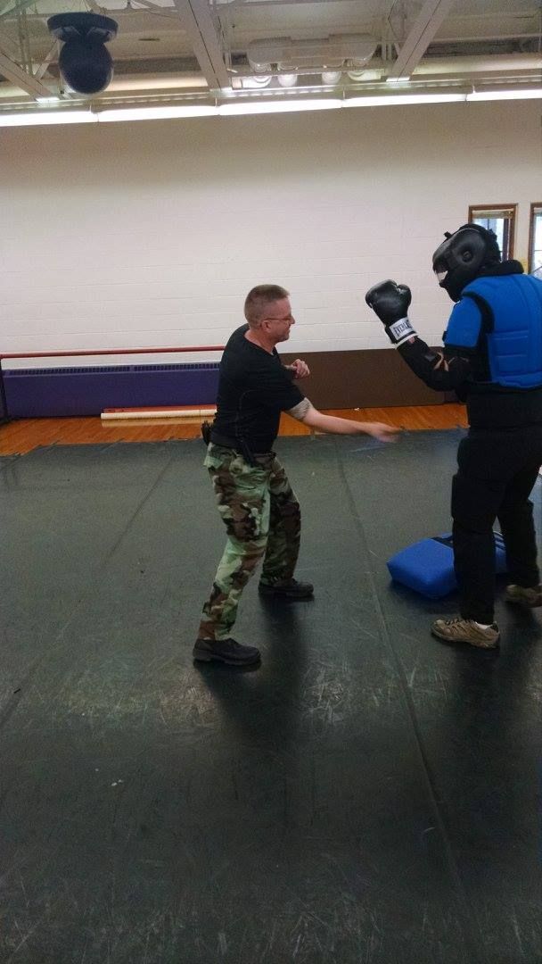 Man in camouflage pants practices martial arts, sparring with a person wearing protective gear in a gym.