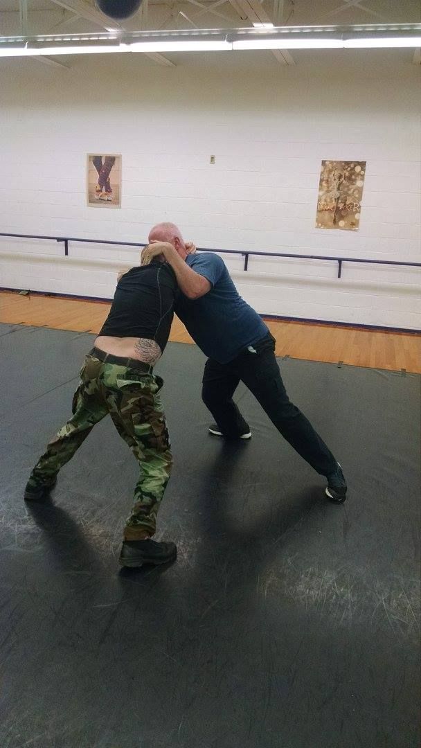 Two men in a wrestling stance on a mat. One wears camouflage pants; the other, a blue shirt, is grabbing the first man's head.