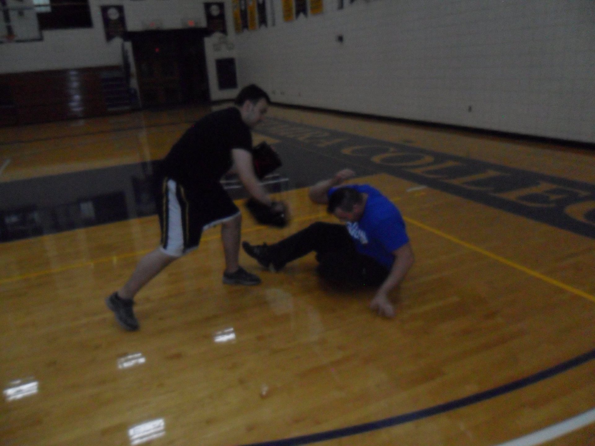 Two men on a wooden basketball court. One, wearing black, crouches with a glove. The other, in blue, is on the floor, possibly knocked down.