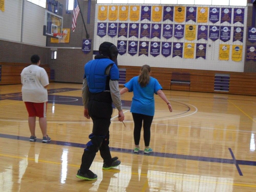A person in fencing gear walks on a gym floor. Two other people stand nearby. Banners hang on the wall.
