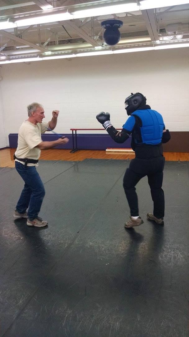 Two men in a gym: one in casual clothes, the other in protective gear, boxing. The man in gear is in a fighting stance.