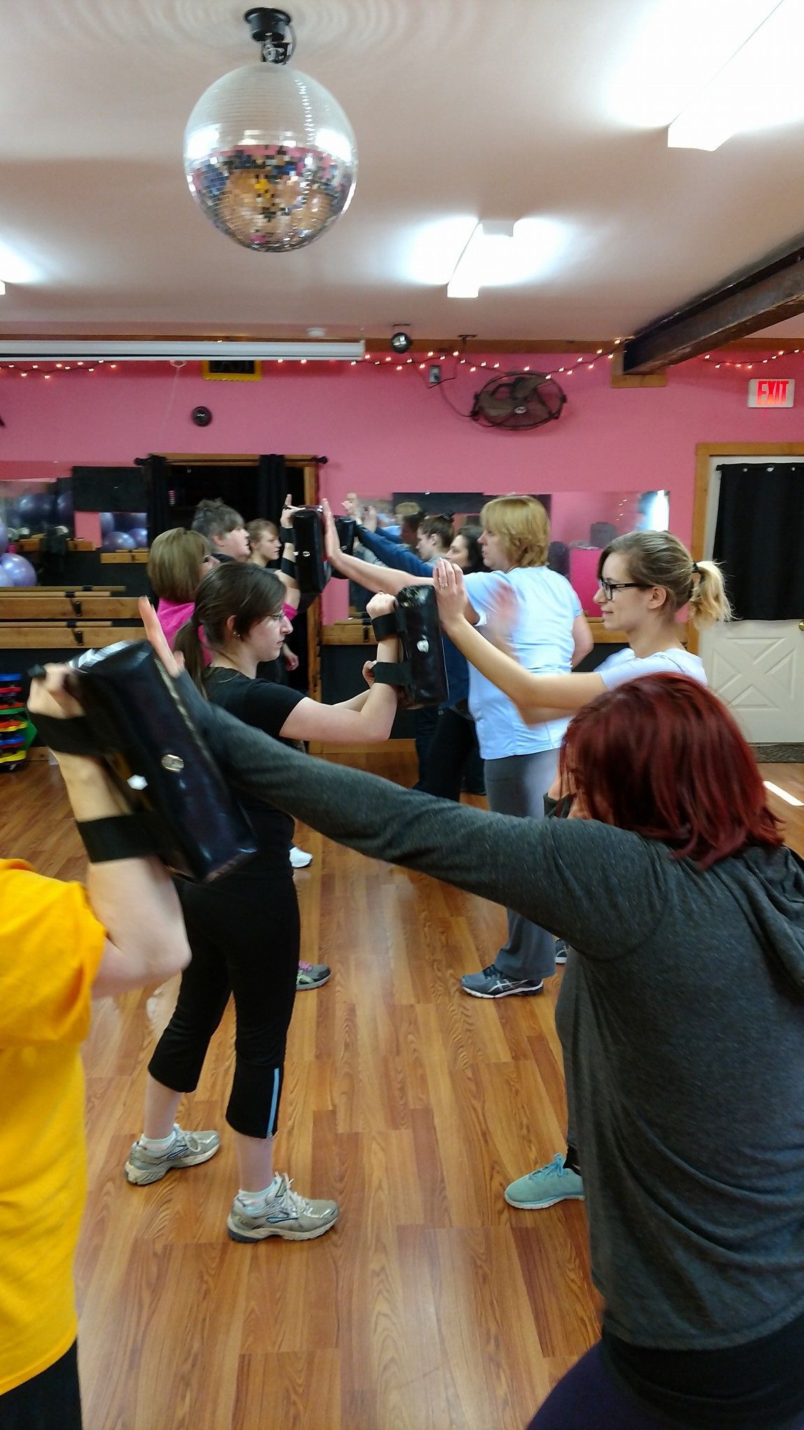 People in a dance studio are participating in a class. Some are holding black pads, others are striking with hands or arms. A disco ball hangs overhead.