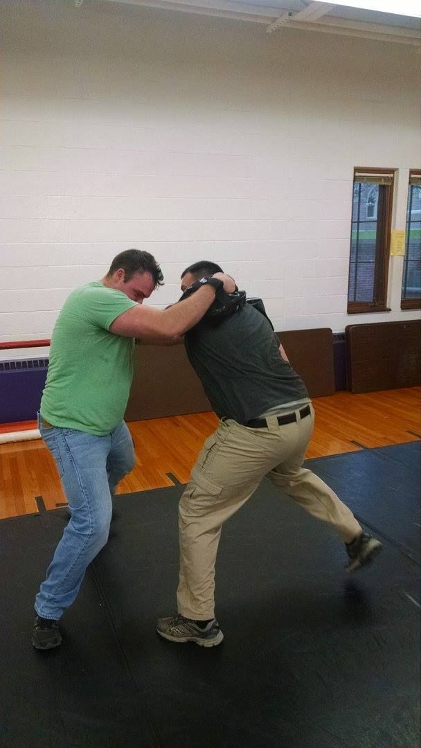 Two men practicing a martial arts move indoors, one in green shirt is grabbing the other’s neck, who is in tan pants and is turning.