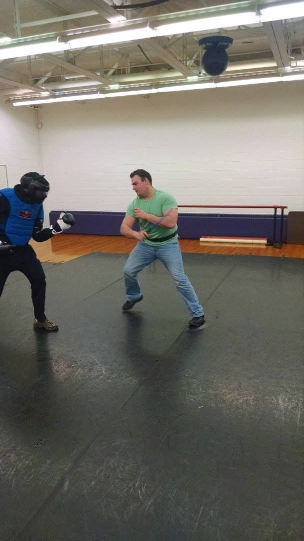 Man in green shirt and jeans in a fighting stance faces person in protective gear in a gym.